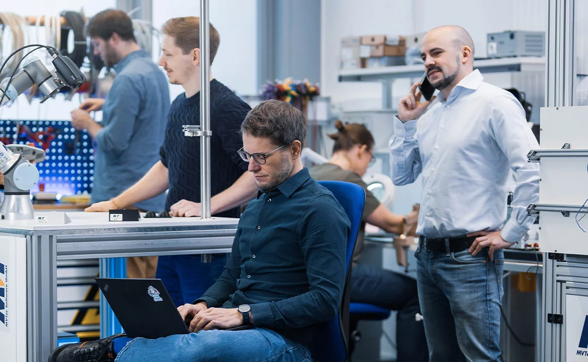 Support team in an engineering lab: one person works on a laptop while others test a robotic arm and hardware at a workbench; a colleague is on the phone.